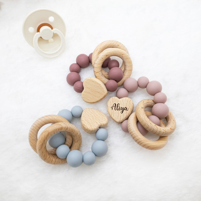 May include: Three wooden teething rings with silicone beads in blue and pink. The rings are circular and have a heart-shaped wooden bead with the name "Aliya" engraved on it.