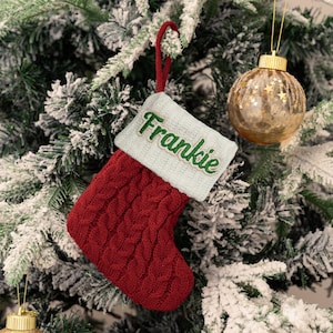 May include: A deep red cable-knit Christmas stocking featuring a light blue cuff with the name Frankie embroidered in green thread. The stocking includes a matching red hanging loop and is displayed against a flocked green pine tree branch.