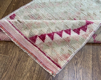 May include: A cream-colored rug with a pink and red geometric pattern. The rug has a fringe border and is folded in half.