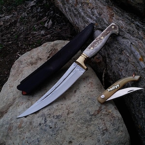 Op de afbeelding: Een gebogen, zilverkleurig mes met een wit en bruin handvat en een gouden metalen accent. Het mes zit in een zwarte leren schede. Een kleiner, vouwmes met een wit en bruin handvat is ook afgebeeld. Het kleinere mes heeft het woord "hediye" op het handvat.