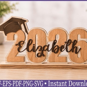 Può includere: Decorazione di laurea in legno con l'anno 2026 e il nome "Elizabeth" in corsivo. Un cappello di laurea è posto sopra il numero 2. La base è marrone chiaro, con marrone più scuro per i numeri, il nome e il cappello.