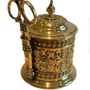 Puede incluir: Un recipiente de metal dorado con un diseño ornamentado y una tapa con bisagras. El recipiente tiene un asa decorativa con dos anillos.