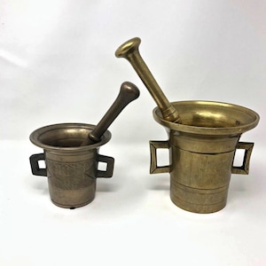 Könnte beinhalten: Zwei antike Mörser und Stößel aus Messing. Die Mörser haben eine zylindrische Form mit Griffen, und die Stößel sind zylindrisch mit abgerundeten Enden. Das Messing hat eine warme, gealterte Patina. Ein Mörser hat geätzte Designs.