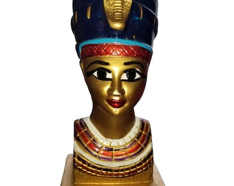 May include: A gold bust of an Egyptian queen wearing a blue and gold headdress with a cobra on top. The bust is on a square base with a black bottom.