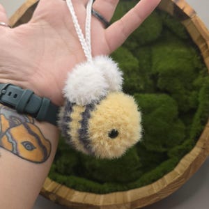 May include: A plush bee ornament with yellow, black, and white fur. The bee has a white fluffy head and is suspended by a white cord. The bee is approximately 2 inches long. The background is a wooden bowl filled with green moss.