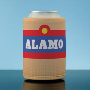 May include: A tan can cooler with the word "ALAMO" in white block letters on a blue stripe, with red stripes above and below. A yellow circle is above the blue stripe. The can cooler is on a reflective surface against a blue background.