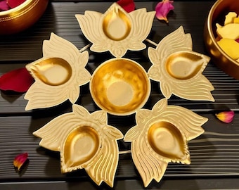 May include: A gold-coloured metal lotus flower candle holder with six individual petal-shaped holders surrounding a central bowl. The candle holder is on a dark surface, with rose petals and small bowls in the background.