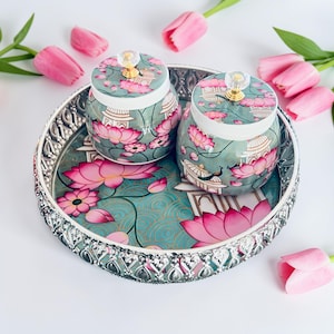 May include: A round silver-toned metal tray with a decorative filigree edge, featuring a teal surface with pink lotus flowers and architectural motifs. Two matching lidded jars with clear crystal-like knobs sit on the tray.