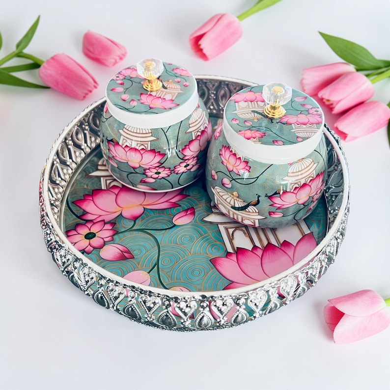 May include: A round silver-toned metal tray with a decorative filigree edge, featuring a teal surface with pink lotus flowers and architectural motifs. Two matching lidded jars with clear crystal-like knobs sit on the tray.