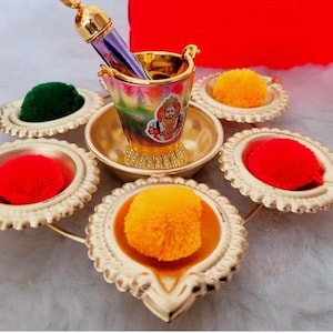 Puede incluir: Un conjunto religioso decorativo con un pequeño cubo colorido con una imagen de deidad, rodeado de pequeños platos dorados que contienen polvos coloridos. Un aplicador dorado está dentro del cubo. El conjunto está dispuesto sobre una superficie clara.