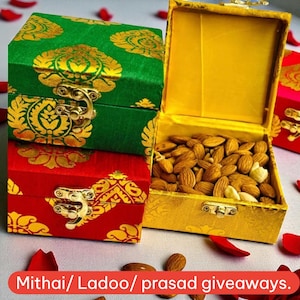 May include: Three decorative boxes, one open and filled with almonds, the others closed. The boxes are green, red, and gold, with ornate gold patterns and clasps. The text "Mithai/ Ladoo/ prasad giveaways" is at the bottom.