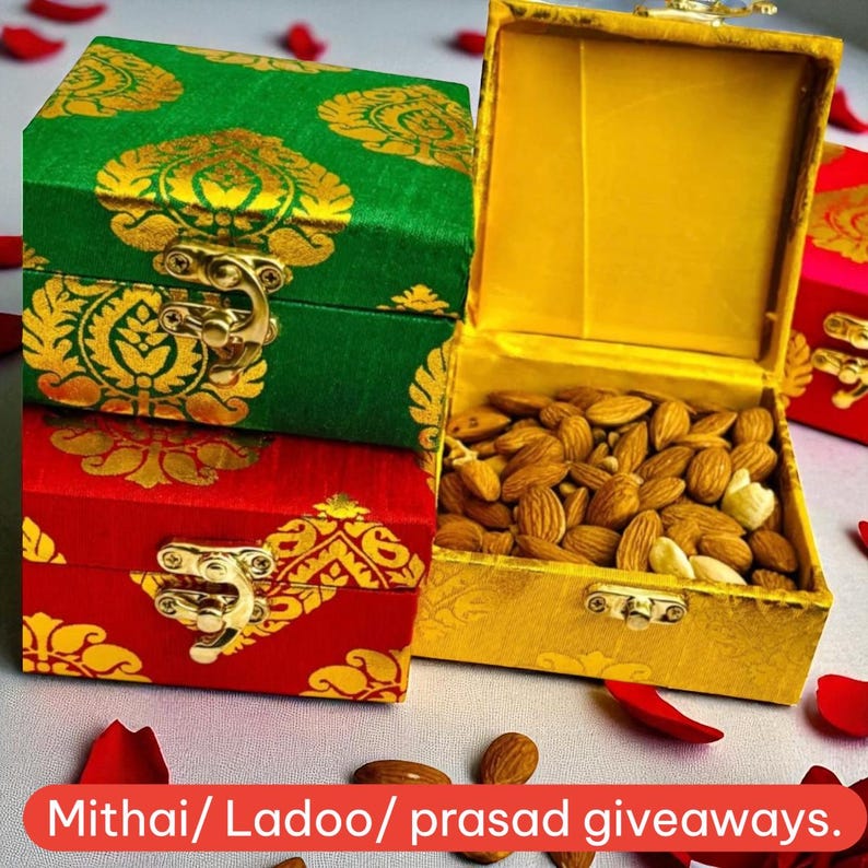 May include: Three decorative boxes, one open and filled with almonds, the others closed. The boxes are green, red, and gold, with ornate gold patterns and clasps. The text "Mithai/ Ladoo/ prasad giveaways" is at the bottom.