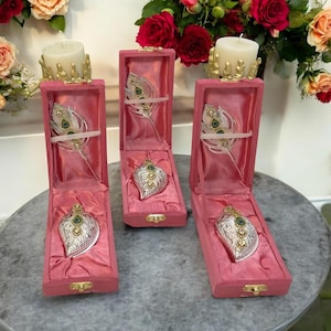Puede incluir: Tres cajas rosas de terciopelo abiertas que muestran colgantes ornamentados plateados en forma de hoja con detalles verdes y dorados. Cada caja también contiene un adorno plateado en forma de pluma. Las cajas están sobre una superficie gris, con flores y velas en el fondo.