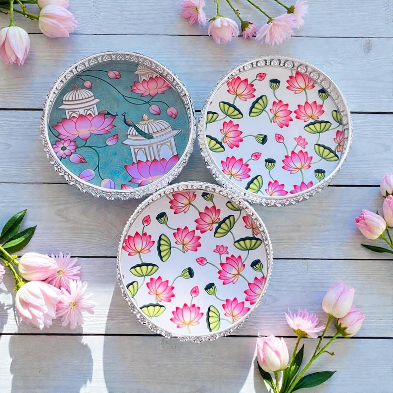 May include: Three round decorative trays with silver-toned scalloped edges. One tray features a teal background with white architectural structures and pink lotus flowers. Two trays feature a white background with a repeating pattern of pink lotus flowers.