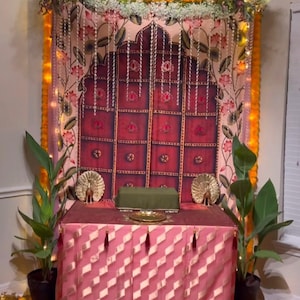 Puede incluir: Un altar decorativo con una falda de mesa rosa y dorada, un tel&oacute;n de fondo granate con dise&ntilde;os florales y una guirnalda de flores. Dos figuras de pavo real doradas flanquean un coj&iacute;n verde sobre la mesa. El altar est&aacute; decorado con guirnaldas de cal&eacute;ndulas.