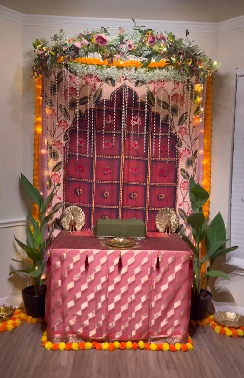Puede incluir: Un altar decorativo con una falda de mesa rosa y dorada, un tel&oacute;n de fondo granate con dise&ntilde;os florales y una guirnalda de flores. Dos figuras de pavo real doradas flanquean un coj&iacute;n verde sobre la mesa. El altar est&aacute; decorado con guirnaldas de cal&eacute;ndulas.