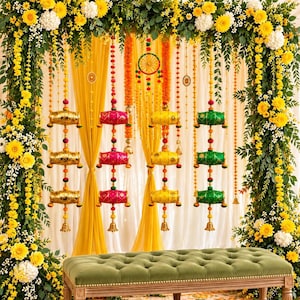 Puede incluir: Un telón de fondo decorativo para eventos con un arco rectangular cubierto de flores amarillas y blancas y follaje verde. Cuelgan hileras de cilindros de tela coloridos en oro, rosa, amarillo y verde, con un banco capitoné verde debajo.