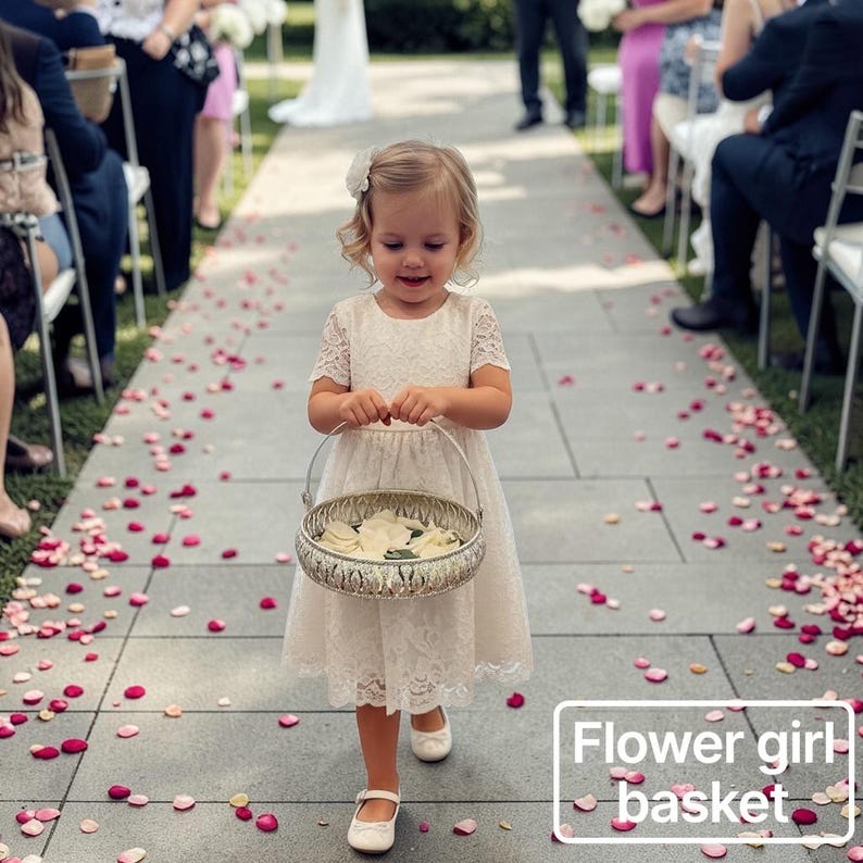 May include: A round, silver-toned metal basket with an intricate filigree pattern and a thin wire handle, filled with white flower petals. The basket is held by a child wearing a cream-colored lace dress and white shoes.