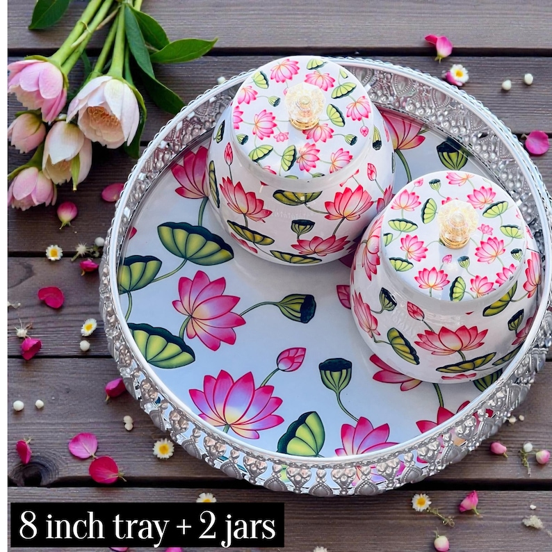 May include: A round 8 inch silver-toned metal tray with a decorative filigree edge, featuring two matching lidded jars. The tray and jars are decorated with a white background and a colorful pattern of pink lotus flowers and green leaves.