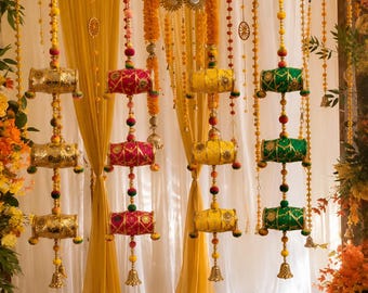 Puede incluir: Una decoración colgante con cuatro hileras verticales de adornos cilíndricos en dorado, rosa, amarillo y verde. Cada hilera incluye pequeños pompones, detalles metálicos y una campanilla en la base, suspendida frente a cortinas de tela amarilla y blanca con arreglos florales.