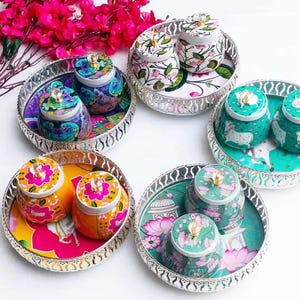 May include: A selection of decorative jars and trays. The jars display vibrant floral and paisley patterns, with jewel-like knobs on the lids. The trays have ornate silver edges and are decorated with matching designs. The colour scheme includes pink, teal, orange, and purple.