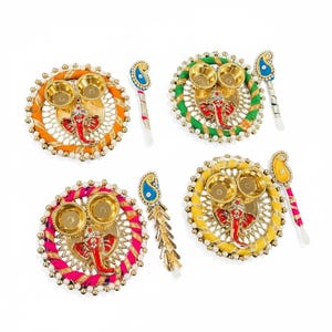 Puede incluir: Cuatro platos decorativos circulares con una figura central de Ganesha roja, dos cuencos pequeños dorados y un borde envuelto en hilo naranja, verde, rosa o amarillo. Cada plato incluye un accesorio a juego en forma de lágrima con cuentas.