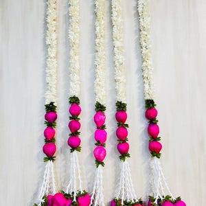 May include: Five vertical garlands with white floral strands and bright pink, heart-shaped ornaments. The garlands are suspended against a light-colored background, creating a visually striking display. The ornaments are clustered at the bottom.
