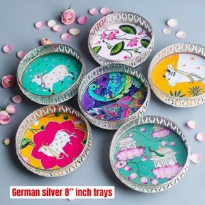 May include: A set of six round German silver 8-inch trays featuring colorful illustrations of cows, peacocks, and lotus flowers, with intricate cut-out metal rims and scattered pink flower petals.