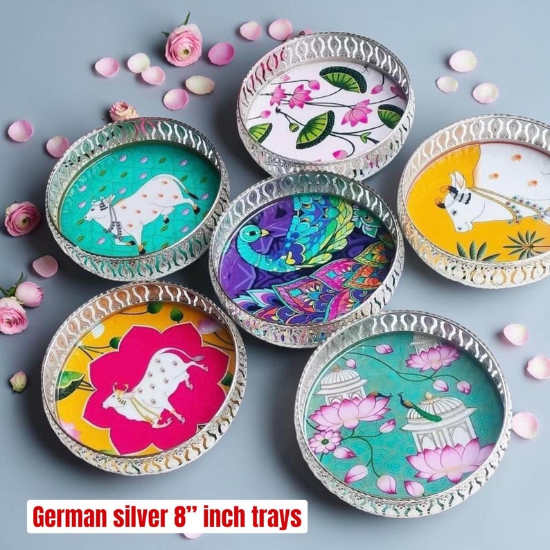 May include: A set of six round German silver 8-inch trays featuring colorful illustrations of cows, peacocks, and lotus flowers, with intricate cut-out metal rims and scattered pink flower petals.