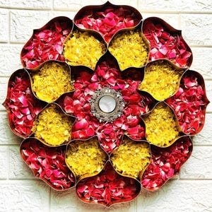 May include: A decorative floral arrangement featuring a central candle holder surrounded by compartments filled with red rose petals and yellow flower petals. The arrangement is set against a white brick wall.