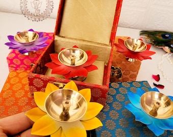 May include: A collection of brass diya oil lamps shaped like lotus flowers with colorful petals in yellow, red, blue, and purple. Each lamp is paired with a small, fabric-covered decorative box featuring intricate gold patterns and metal latches.