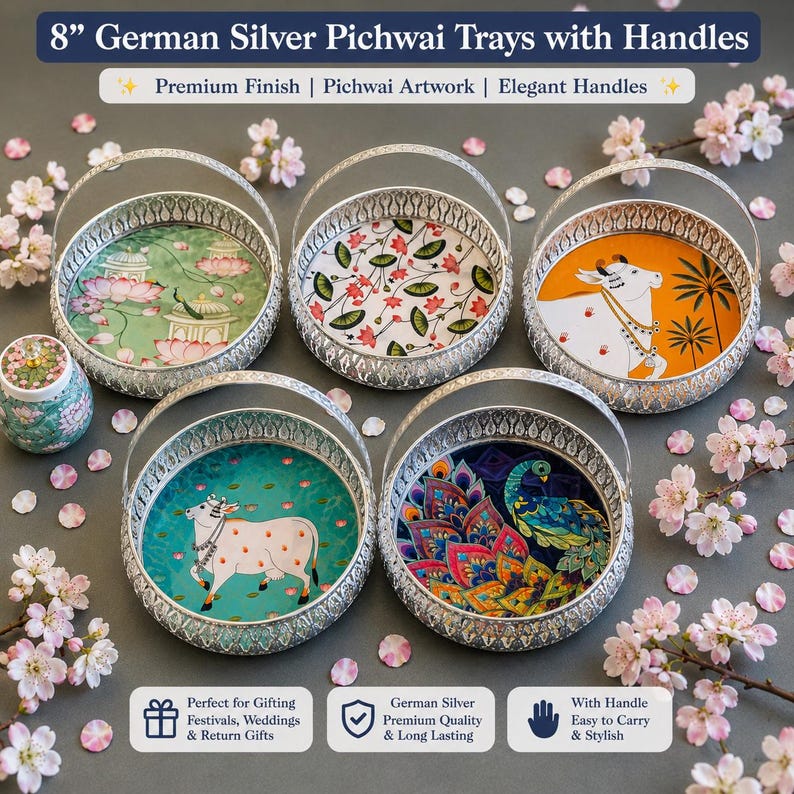 May include: Five round 8-inch German silver trays with arched handles, each featuring distinct colorful Pichwai-style illustrations including cows, peacocks, and lotus flowers, arranged with scattered pink cherry blossom petals.