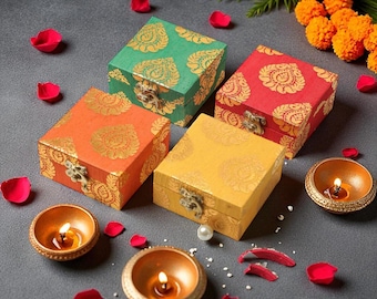 May include: Four colorful, square boxes with ornate gold designs and metal clasps. The boxes are in shades of green, red, orange, and yellow. Small, lit candles in gold-colored bowls and scattered rose petals surround the boxes.