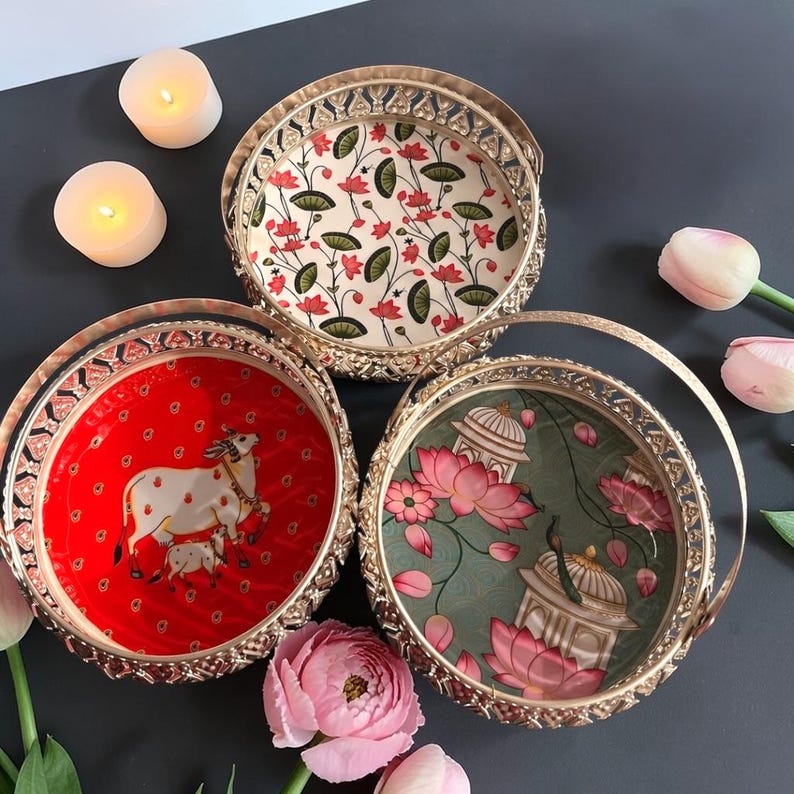 May include: Three round gold-toned metal baskets with decorative cut-out rims and handles. One basket features a red interior with a white cow and calf illustration, another has a cream interior with pink lotus flowers, and the third has a green interior with pink lotus flowers and architectural structures.