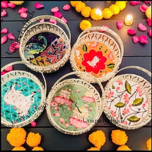 May include: Five round silver-toned metal baskets with arched handles, each featuring a unique decorative base insert. Designs include a peacock, a white cow on orange, a white cow on turquoise, a lotus flower scene, and a floral pattern.