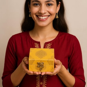 May include: A smiling person holds a yellow gift box with a decorative pattern and a metal clasp. The box is labeled "Handmade Gift Packaging" in gold lettering. The person is wearing a red top.