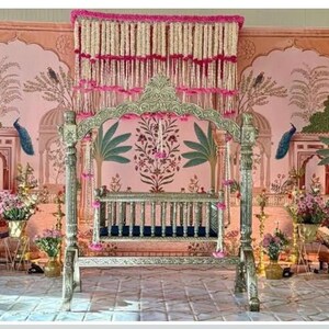 May include: A silver-toned metal swing with a dark blue seat, positioned in front of a pink backdrop featuring illustrations of arches, palm trees, and peacocks. The display is decorated with hanging white and pink floral garlands and gold lamps.