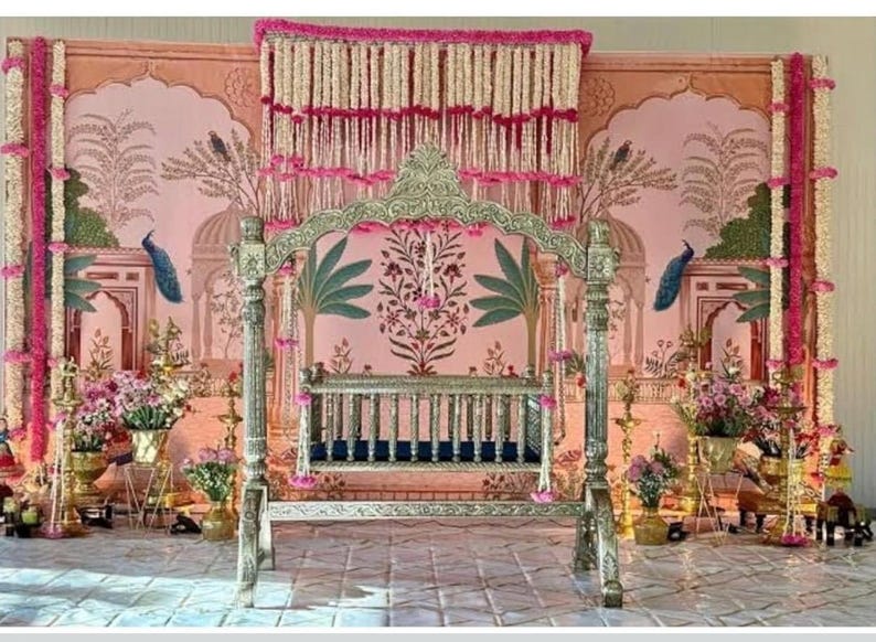 May include: A silver-toned metal swing with a dark blue seat, positioned in front of a pink backdrop featuring illustrations of arches, palm trees, and peacocks. The display is decorated with hanging white and pink floral garlands and gold lamps.