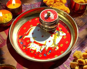 May include: A decorative brass plate with a red background featuring a white cow and calf design. A small, ornate container sits in the center. The plate is surrounded by small bowls and lit candles, suggesting a cultural or religious theme.