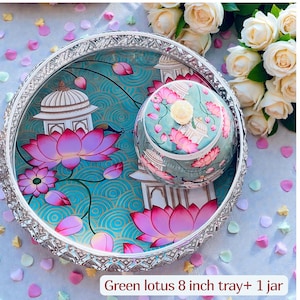 May include: A round silver-toned tray and a matching lidded jar featuring a teal background with pink lotus flowers and white architectural structures. The set includes an 8 inch tray and one jar, both decorated with a repeating pattern of blossoms.