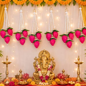 May include: A golden Ganesha statue sits centrally, adorned with a white and pink flower garland. Six hanging decorations feature strings of white faux pearls ending in clusters of three bright pink lotus buds with green leaves.