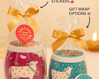 May include: Two Diwali gift hampers with gold bows, one in pink and the other in teal, each with a cow illustration. The pink container has a red "Happy Diwali" sticker. The image includes text: "Customize Your Diwali Gift Hampers."