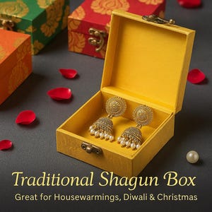 May include: A pair of gold-colored earrings with pearl accents rests inside an open yellow box. The box is labeled "Traditional Shagun Box" and is surrounded by red rose petals. Other decorative boxes are in the background.
