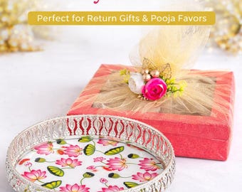 May include: A round silver-toned tray with a white base featuring a pink and green lotus flower pattern, accompanied by a square red gift box topped with a gold mesh bow and artificial flowers. The text reads Lotus Pichwai Tray Gift Set.