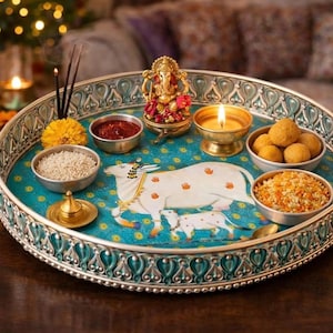 May include: A round, silver-toned metal tray with an intricate teal and silver border. The center features a painted white cow and calf on a teal background. It holds small metal bowls containing rice, red powder, and yellow grains, plus a golden Ganesha figurine and a lit candle.