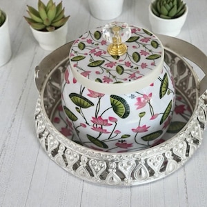 May include: A white ceramic jar with a pink lotus flower and green leaf pattern, topped with a clear crystal knob and gold base. The jar sits inside a round, silver-toned metal tray featuring an intricate, openwork filigree border design.