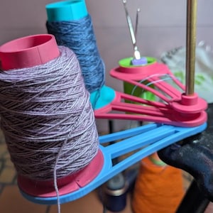 May include: A close-up of a yarn winder with several spools of yarn in various colors. The yarn spools are pink, blue, and orange. The winder is black and has a blue and pink yarn holder.