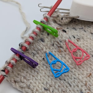 May include: Four colorful stitch markers in the shape of gnomes are placed on a beige knitted fabric. The markers are purple, green, blue, and pink. A red knitting needle is in the background.
