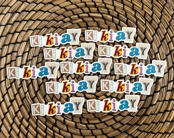 May include: A set of 10 colorful stickers with the text "ki ki ay" repeated on each sticker. The stickers are arranged in a circle on a woven brown background.