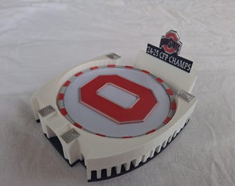 May include: A white and red decorative stadium-shaped item with a large red letter "O" in the center. The item has a sign that reads "Ohio State 24-25 CFP CHAMPS".